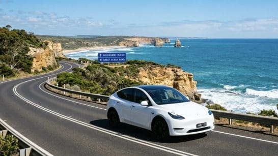 White electric car on a winding coastal road with rocky cliffs and the blue ocean; road signs indicate Melbourne 150 km and the Great Ocean Road.